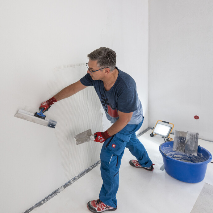 Heimwerker zieht im Raum einen weißen, feinen Oberputz eben mit einem Flächenspachtel ab. Um Unebenheiten zu sehen, wird die Wand von einem Baustrahler angestrahlt.