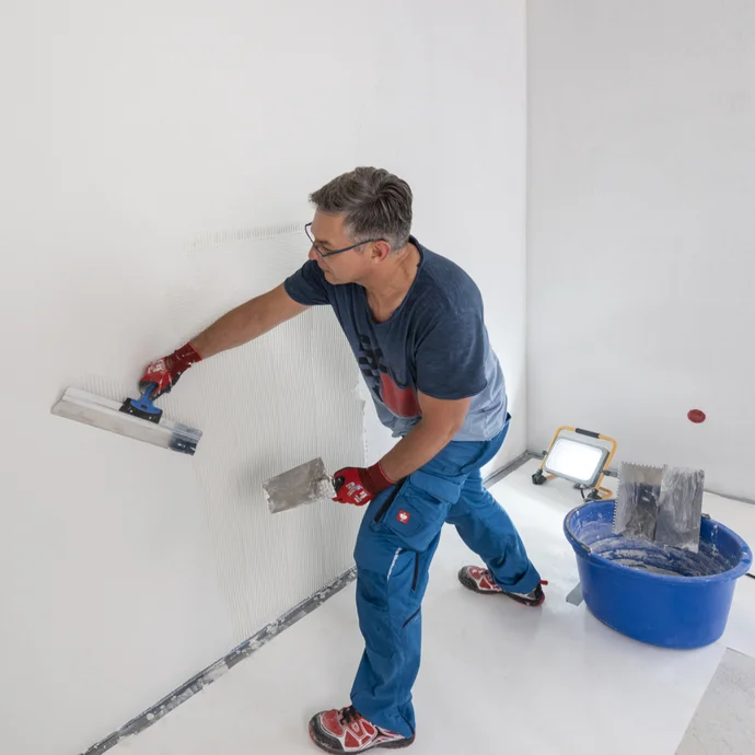 Heimwerker zieht im Raum einen weißen, feinen Oberputz eben mit einem Flächenspachtel ab. Um Unebenheiten zu sehen, wird die Wand von einem Baustrahler angestrahlt.