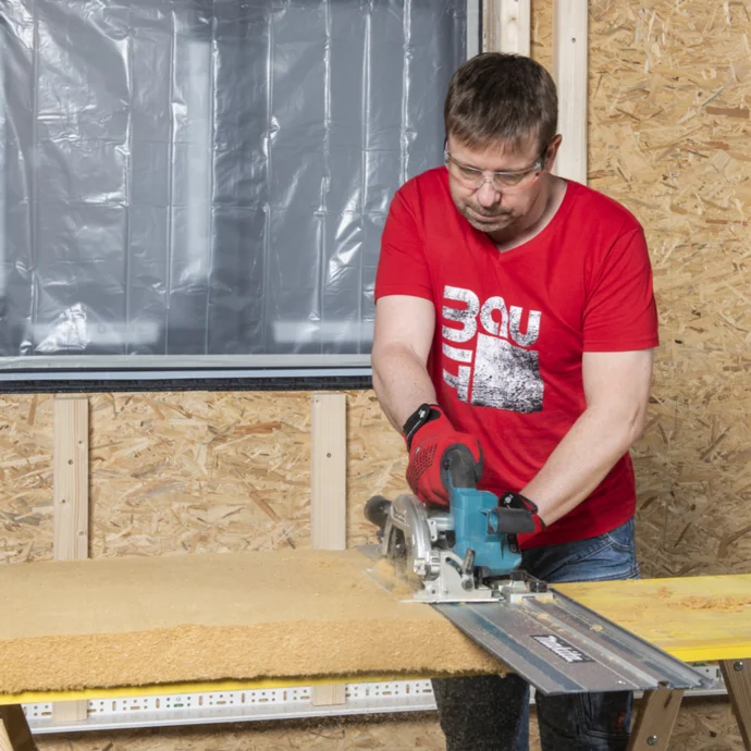Mann beim Schneiden von Holz mit Kreissäge im Innenausbau, trägt rotes Baumit-Shirt vor OSB-Plattenwand.
