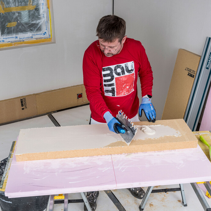 Heimwerker trägt Baumit Klebemörtel mit einer Zahnkelle als Pressspachtelung dünn und vollflächig auf die Klebeseite der Holzfaserdämmplatte auf – optimale Haftung zwischen Kleber und Dämmplatte für die Innendämmung, Baumit Selbermachen Anleitung.
