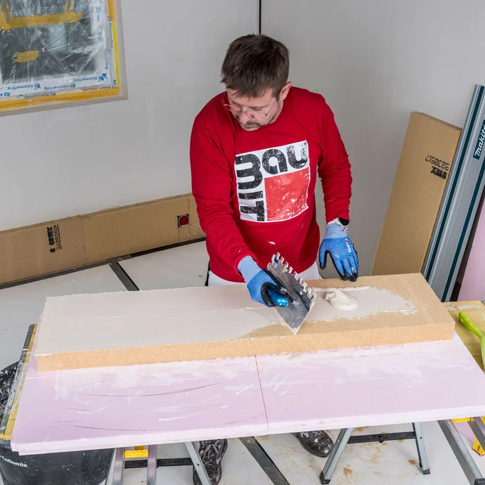 Heimwerker trägt Baumit Klebemörtel mit einer Zahnkelle als Pressspachtelung dünn und vollflächig auf die Klebeseite der Holzfaserdämmplatte auf – optimale Haftung zwischen Kleber und Dämmplatte für die Innendämmung, Baumit Selbermachen Anleitung.