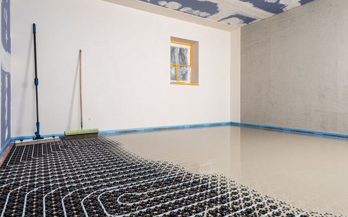 Foto eines Innenraumes: Bodenheizung ist verlegt, Schicht Ausgleichsmasse wird eingearbeitet. In der rechten Ecke stehen Werkzeuge - eine Spachtel und eine Stachelwalze.