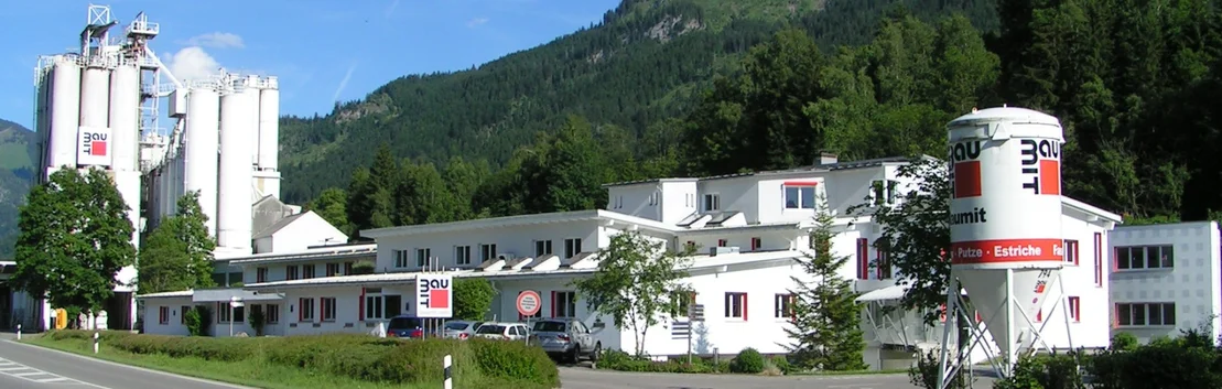 Fabrikgeb&auml;ude von Baumit in idyllischer Berglandschaft, umgeben von Pflanzen und Silos im Hintergrund.