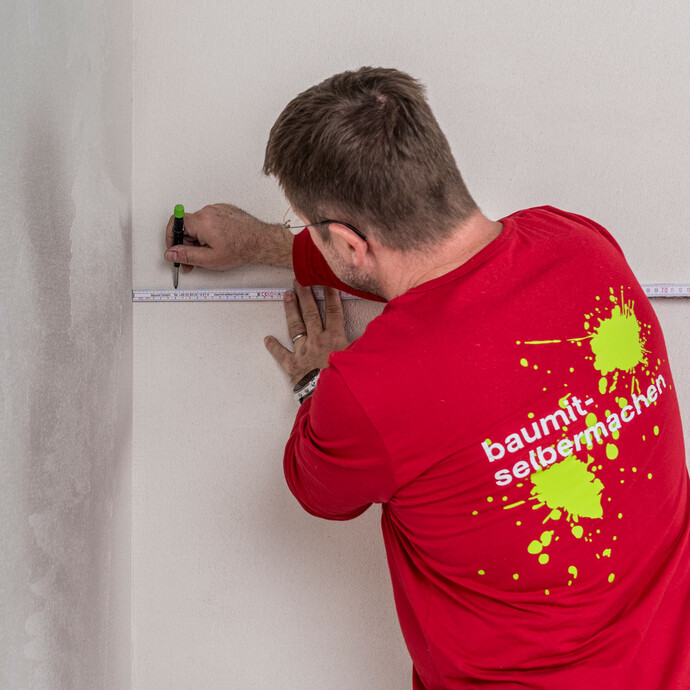 Heimwerker markiert mit Zollstock und Stift die richtige Position für das Baumit Fugendichtband im Stoßbereich an der Innenwand – Markierungspunkte setzen für eine bündige Verklebung der Holzfaserdämmplatten, Baumit Selbermachen Anleitung.