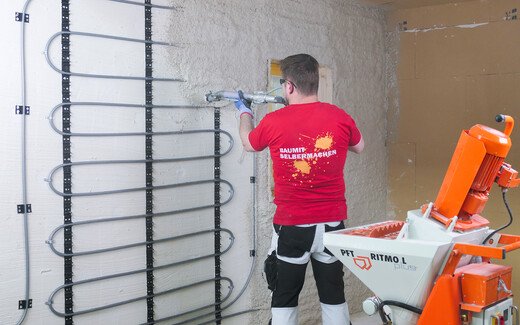 Heimwerker verputzt Wand mit Putzmaschine vor installierter Wandheizung – maschineller Putzauftrag im Innenbereich.