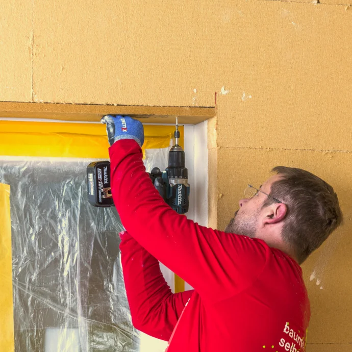 Person montiert mit einer Bohrmaschine ein Holzfenster, umrahmt von gelber Schutzfolie, in einer dämmstoffverkleideten Wand.