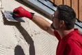 Heimwerker trägt Außenputz auf Fassade auf. Pazifisches Rot am Handschuh für sicheren Grip. Ideal für DIY-Projekte.