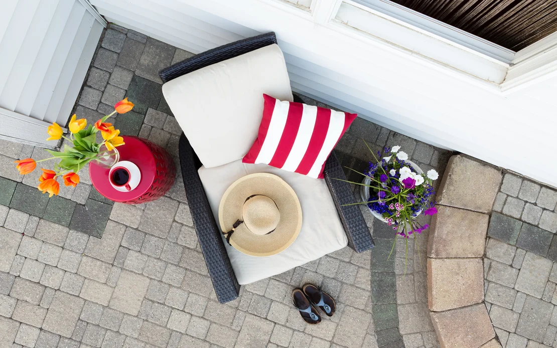Gem&uuml;tlicher Outdoor-Loungesessel auf gepflasterter Terrasse mit Dekokissen und Blumenarrangement, ideal f&uuml;r Heimwerkerprojekte.
