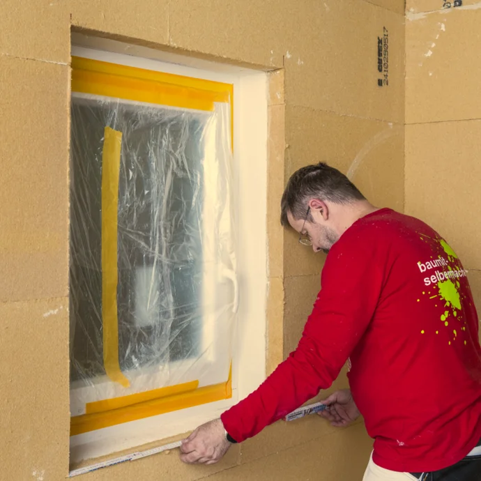 Handwerker beim Abkleben eines Fensterrahmens mit Folie und Klebeband zur Vorbereitung von Putzarbeiten.