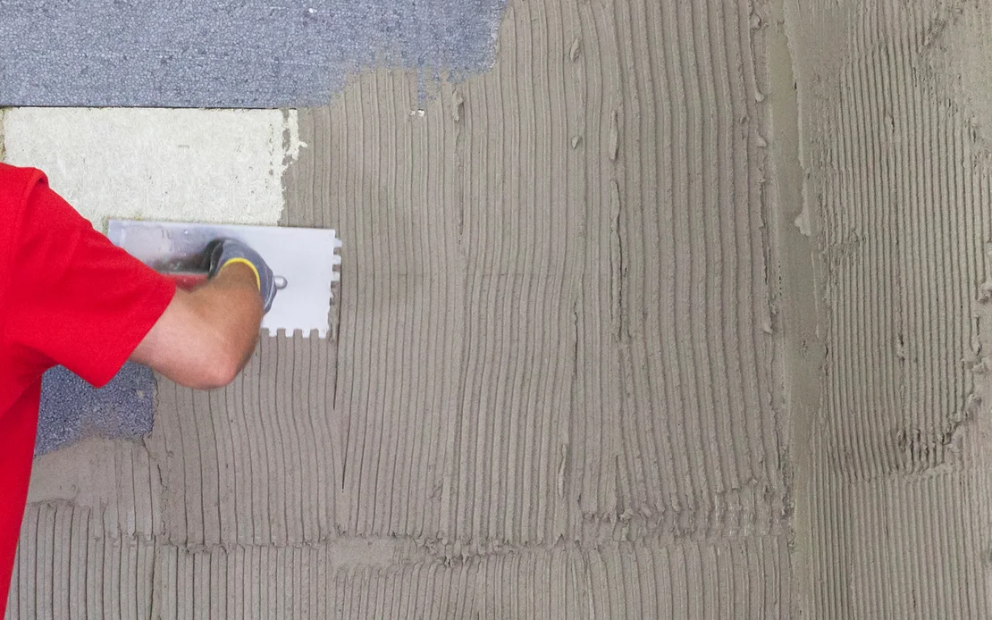 Person trägt Kleber mit Zahnspachtel auf Wand auf, für Fliesenverlegung vorbereitet.