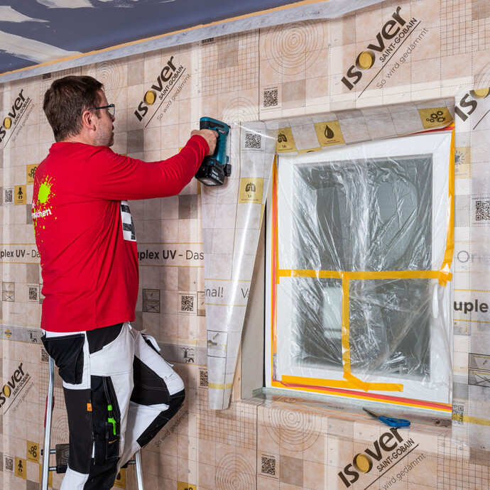 Heimwerker richtet das zugeschnittene Folienstück der Dampfbremse an der Laibung des Fensters aus und tackert es an der Holzkonstruktion fest. Baumit Anleitung zum Selbermachen: Dampfbremse in einer Fensterlaibung richtig einbauen.