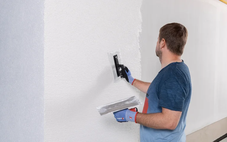 Mann trägt mit Spachtel Putz auf Innenwand auf, Schritt-für-Schritt-Anleitung zur Wandrenovierung mit Baumit-Produkten.