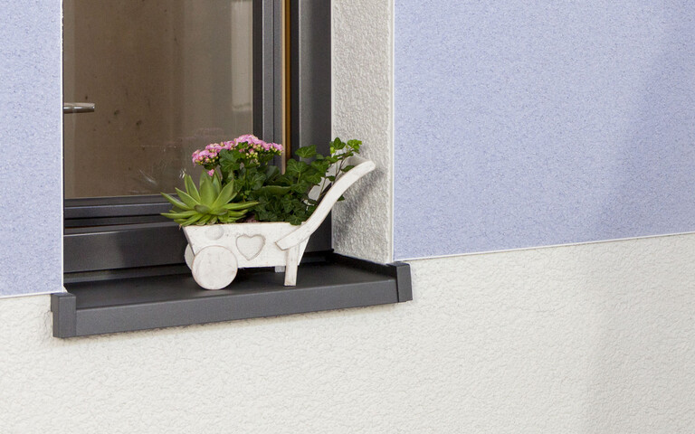 Dekorativ verputzte Fassade mit blauer Kratzputzfläche und Blumen am Fensterbrett.