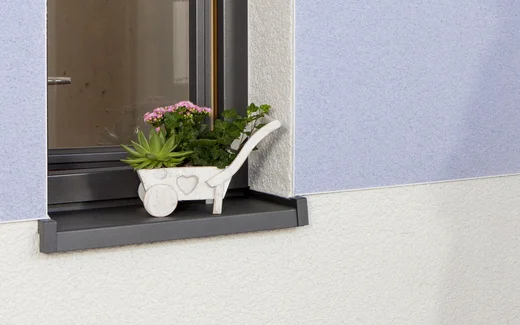 Dekorativ verputzte Fassade mit blauer Kratzputzfläche und Blumen am Fensterbrett.