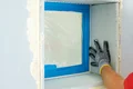 Heimwerker mit Handschuh glättet Wände an einer Fensteröffnung, vorbereitet für Verputzarbeiten mit blauer Schutzfolie.