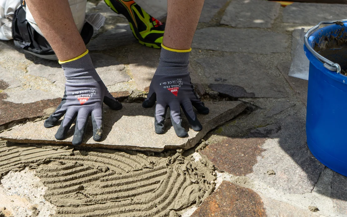 Handwerker dr&uuml;ckt eine Natursteinplatte mit Handschuhen in frischen Kleber auf einem gepflasterten Boden fest.
