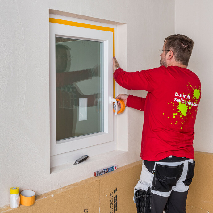 Heimwerker klebt das Fenster mit einem Klebeband mit etwas Abstand zum Mauerwerk ab, um das Fenster vor Schmutz zu schützen.