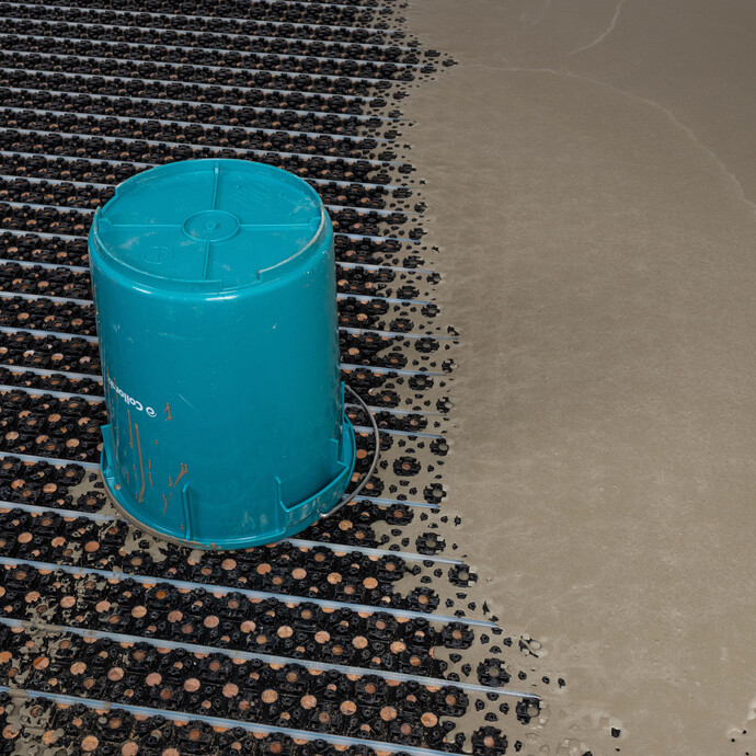 Leerer, blauer Eimer steht umgedreht auf der neu eingebauten Fußbodenheizung mit Noppenplatte. Rechts davon ist die frisch ausgeleerte Ausgleichsmasse erkennbar.