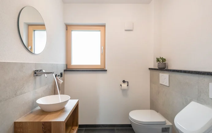 Modernes Gäste-WC mit weißem Waschbecken mit fließendem Wasser auf Sideboard aus Holz inszeniert mit edlen grauen Wandplatten und wohngesundem Edelputz InStyle Edelweiß in feiner, gefilzter Struktur.