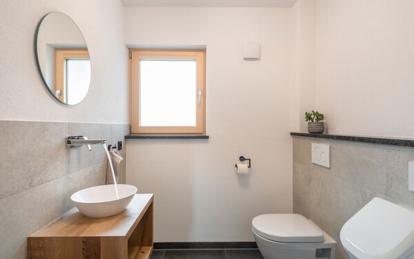 Modernes Gäste-WC mit weißem Waschbecken mit fließendem Wasser auf Sideboard aus Holz inszeniert mit edlen grauen Wandplatten und wohngesundem Edelputz InStyle Edelweiß in feiner, gefilzter Struktur. 