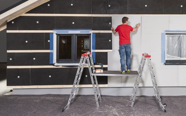 Heimwerker installiert Fassadendämmplatten an einem Haus mit Hilfe von zwei Leitern und einem Arbeitsbrett.