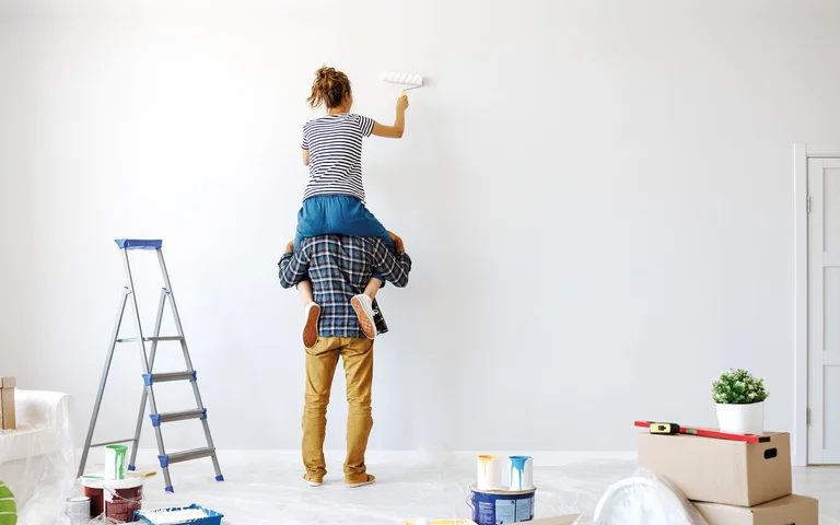Paar beim Streichen einer Wand, Frau auf Leiter, Baumaterialien im Vordergrund, DIY-Heimwerkerprojekt.