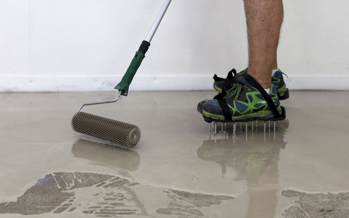 Mann steht mit Nagelsohlen in Ausgleichsmasse und entlüftet diese mit einer Stachelwalze.