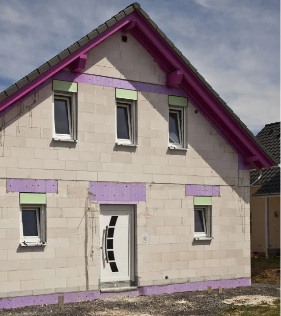 Fassade eines Rohbaus mit unverputzten Mauersteinen, lila Akzenten und weißer Eingangstür, bereit zur Fertigstellung.