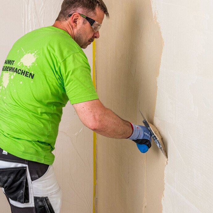 Heimwerker trägt Baumit Kalkin Kalkspachtel multifine als Grundspachtelung mit der Glättkelle auf die Innenwand auf – minimale Unebenheiten begradigen für eine glatte Oberfläche, Baumit Selbermachen Anleitung.