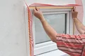 Person bringt Fensterdichtband an weißem Fensterrahmen an, Schritt in Heimwerkerprojekt zur energetischen Sanierung.