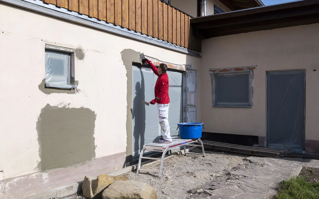 Mann steht auf einem Ger&uuml;st und verputzt das Kantenprofil und die Hohlstellen oberhalb der Terrassent&uuml;re. Daneben steht ein blauer M&ouml;rtelk&uuml;bel mit den Fassadenspachtel.