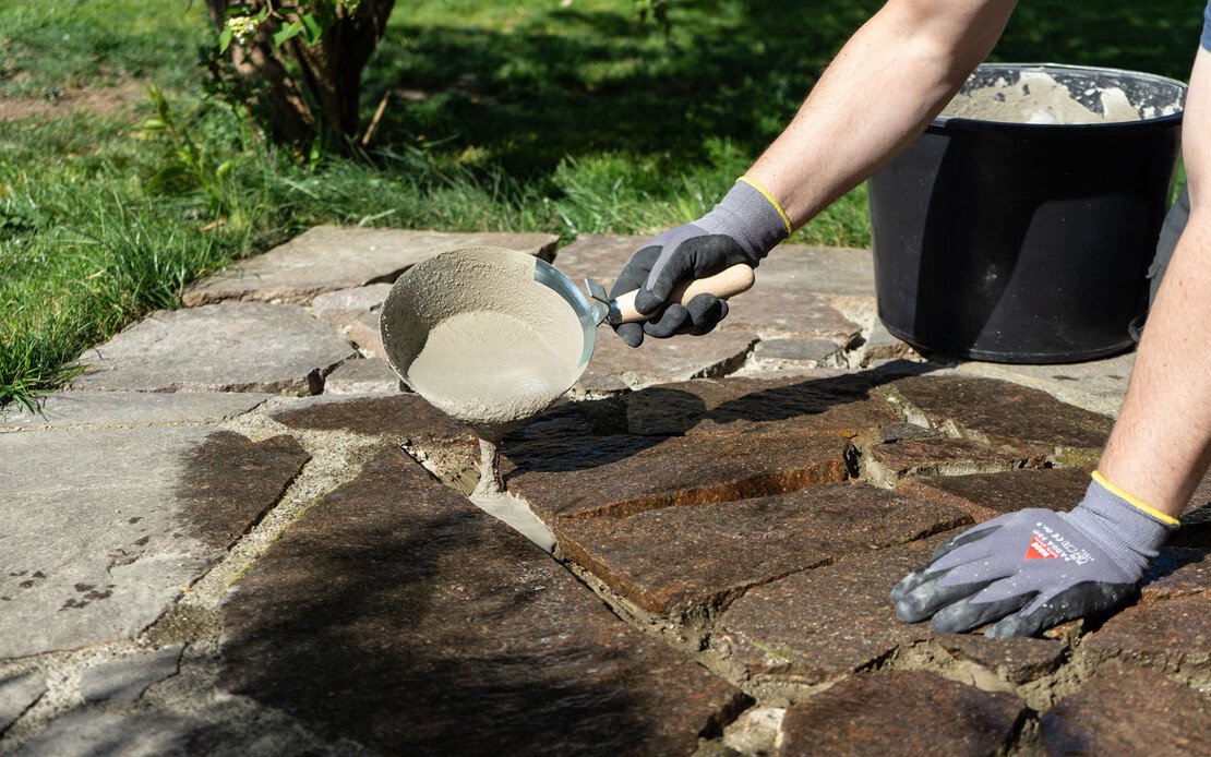 Person gie&szlig;t Fugenm&ouml;rtel in die Fugen der Natursteinplatten im Au&szlig;enbereich.