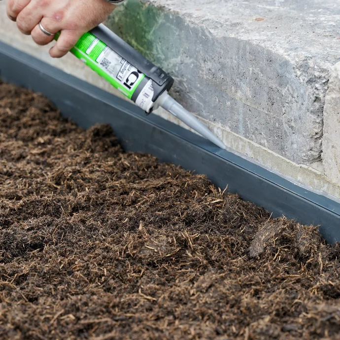 Handwerker dichtet das Abschlussprofil mit einer grünen Kartusche Silikon ab zum Schutz vor eindringender Feuchtigkeit. Die helle Silikonfuge ist auf den gräulichen Stein der Gartenmauer abgestimmt.