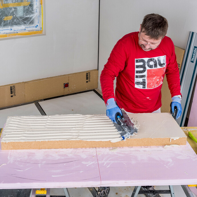Heimwerker trägt Baumit Klebemörtel vollflächig auf Holzfaserdämmplatte auf und zahnt die Fläche mit einer Zahnkelle auf – vollflächige Verklebung der Innendämmung ohne Hohlräume sicherstellen, Baumit Selbermachen Anleitung.
