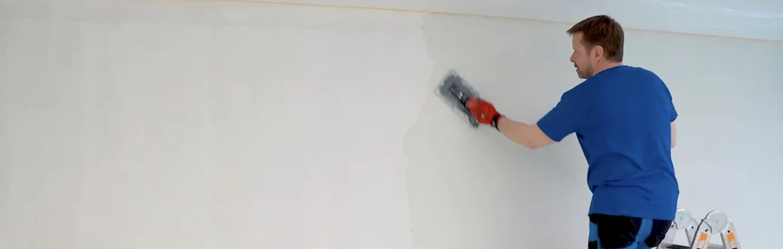 Heimwerker trägt mit einer Kelle Putz auf eine Innenwand auf, um sie für das Streichen vorzubereiten.