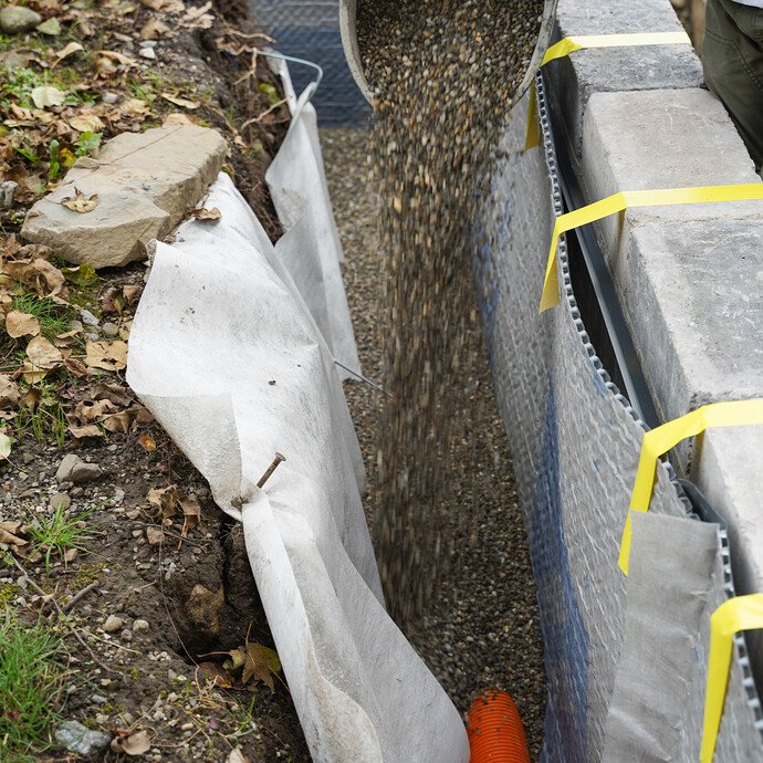 Detailaufnahme: Eimer mit Kies wird in die Baugrube auf das verlegte Drainagerohr gelegt.