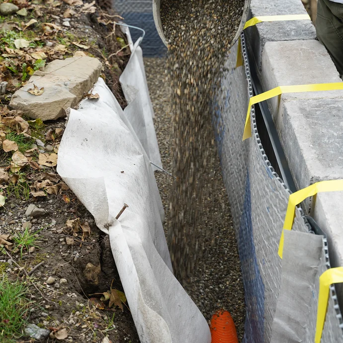 Detailaufnahme: Eimer mit Kies wird in die Baugrube auf das verlegte Drainagerohr gelegt.