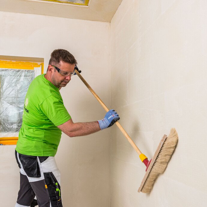 Heimwerker kehrt Innenwand mit einem Besen ab, um Staub und lose Partikel zu entfernen – Untergrundvorbereitung vor dem Auftragen von Baumit Kalkin Kalkspachtel.