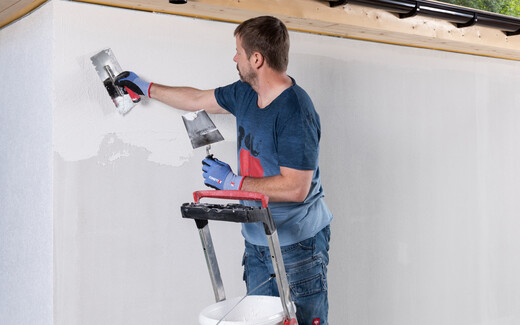 Person steht auf Leiter und trägt Strukturputz mit einer Glättkelle an einer Wand auf.