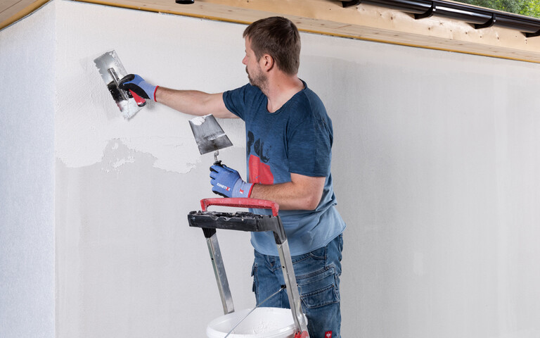 Person steht auf Leiter und trägt Strukturputz mit einer Glättkelle an einer Wand auf.