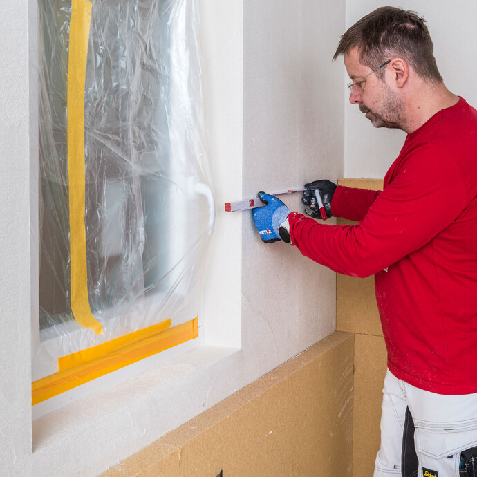 Heimwerker misst den L-Schnitt am Fenster aus. So passt die Holzfaserdämmplatte direkt unter das Fenster. Innendämmung Anleitung Baumit Selbermachen.