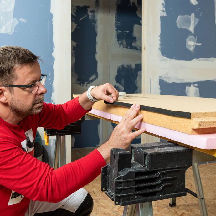 Heimwerker klebt Baumit Fugendichtband auf die Unterseite einer Holzfaserdämmplatte – Vorbereitung für die erste Plattenreihe bei der Innendämmung mit Holztragkonstruktion.