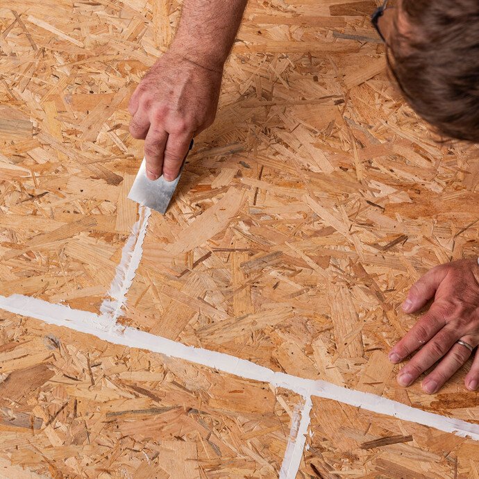 Person zieht den aufgetragenen Montagekleber, der in die Stoßfugen der Spanplatten aufgetragen wurde, oberflächenbündig mit einer Spachtel ab.
