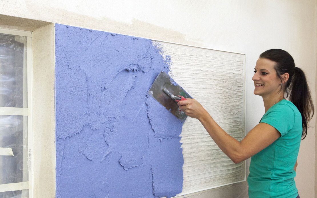 Frau trägt blauen Kratzputz mit einer Glättkelle auf die vorbereitete Fassade auf.