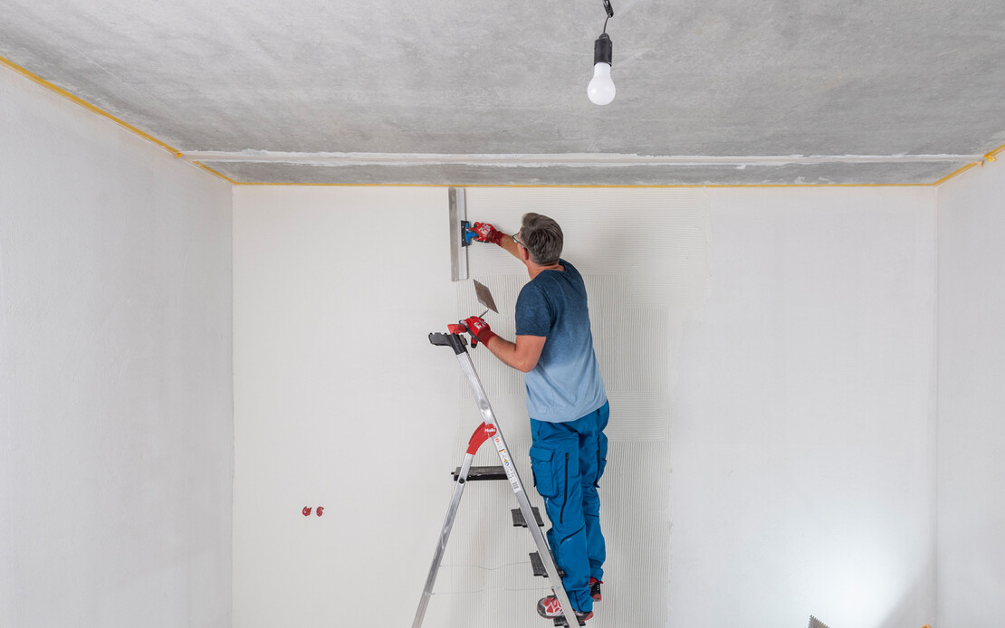 Mann steht im Raum auf einer Leiter und zieht den weißen, feinen Putz mit einem Flächenspachtel ab. Um Unebenheiten zu sehen, wird die Wand von einem Baustrahler angestrahlt.