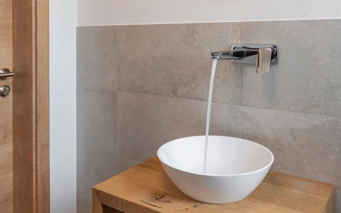 Modernes, weißes Waschbecken mit fließendem Wasser auf Sideboard aus Holz inszeniert mit edlen grauen Wandplatten mit wohngesundem Edelputz in gefilzter Struktur.