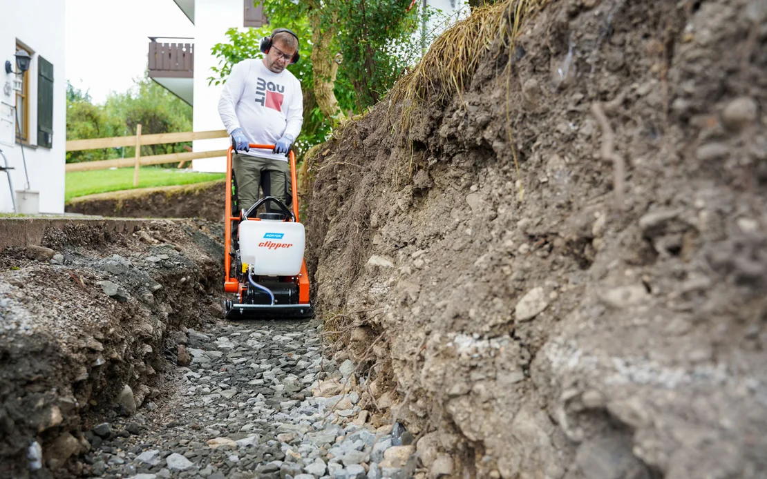 Heimwerker verdichtet den Kiesuntergrund im Garten mit einer R&uuml;ttelplatte f&uuml;r ein stabiles Fundament.