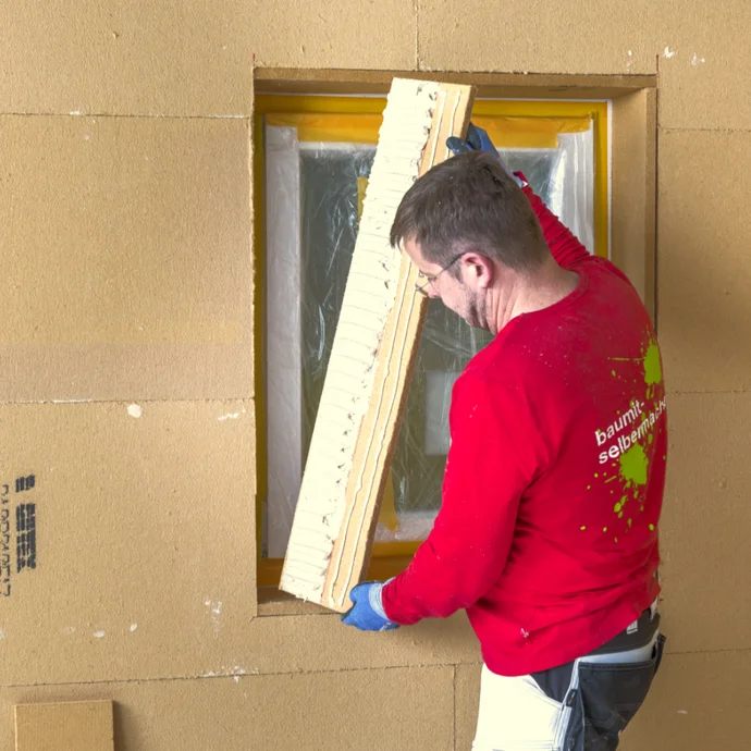 Heimwerker beim Einbau einer Dämmplatte in die Wand eines Rohbaus mit Fenster, Arbeitskleidung und Werkzeug.
