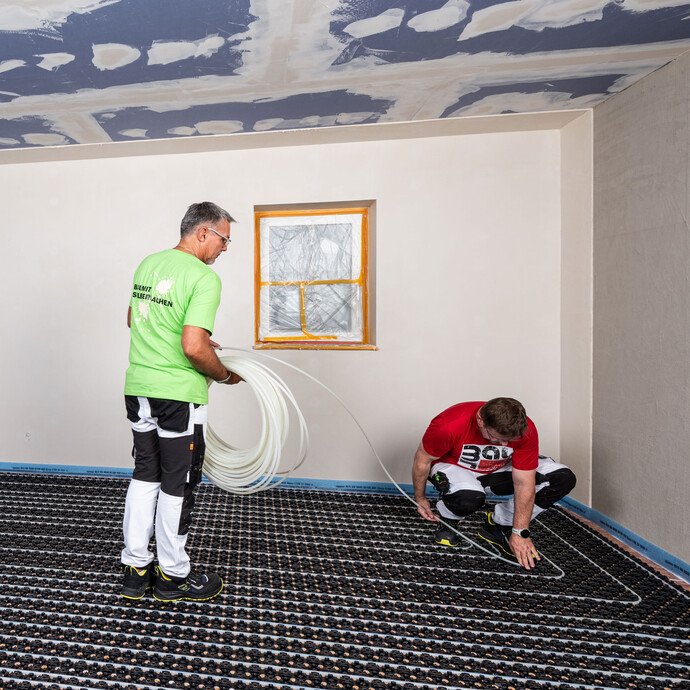 Zwei Handwerker verlegen den Heizungsschlauch der Minitec Fußbodenheizung in die Noppenplatten. Die erste Person hält den Schlauch, die zweite Person verlegt den Heizungsschlauch.