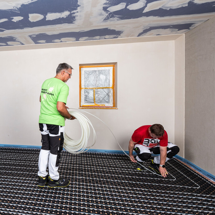 Zwei Handwerker verlegen den Heizungsschlauch der Minitec Fußbodenheizung in die Noppenplatten. Die erste Person hält den Schlauch, die zweite Person verlegt den Heizungsschlauch.